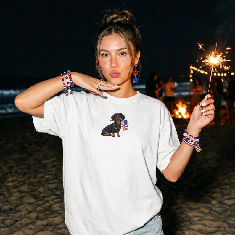 Op de afbeelding: Wit T-shirt met een afbeelding van een teckel die een Amerikaanse vlag vasthoudt. De hond is zwart en bruin. De persoon draagt armbanden en oorbellen met rode, witte en blauwe sterren. Een brandende sterretjes is in de hand van de persoon.