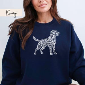 May include: Navy blue sweatshirt featuring a white floral patterned dog silhouette. The word "Navy" is written in cursive in the upper left corner. The sweatshirt is a solid navy color.