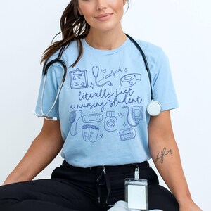 May include: A light blue t-shirt with a graphic of medical supplies and the text "literally just a nursing student".