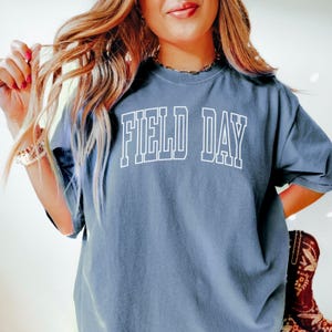 May include: A dusty blue t-shirt with the words "FIELD DAY" in white block letters. The shirt has a relaxed fit and short sleeves. The text is outlined in white, creating a bold contrast against the shirt's color.
