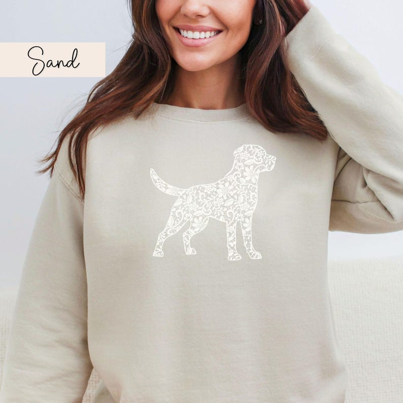 May include: A sand-colored sweatshirt featuring a white floral dog silhouette. The word "Sand" is written in cursive in the upper left corner. The sweatshirt has long sleeves and a crew neck.