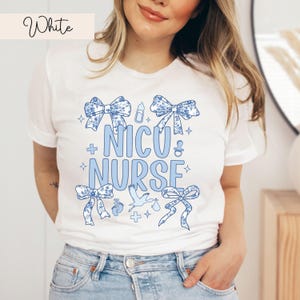 May include: White t-shirt with a blue and white floral design. The shirt has the text "NICU NURSE" with a baby bottle, a stork, and baby footprints.