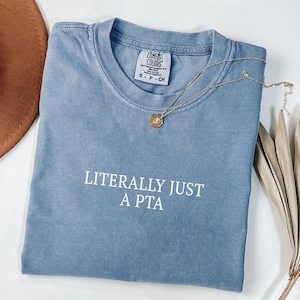 May include: A blue short-sleeved t-shirt with the text "LITERALLY JUST A PTA" in white. A gold necklace with a pendant is draped across the shirt. The shirt is folded on a white surface.