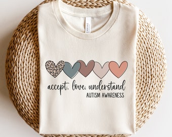 May include: A beige t-shirt with a row of five colorful hearts and the text "accept. love. understand. AUTISM AWARENESS".