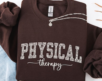 May include: A dark brown sweatshirt with the words "PHYSICAL therapy" printed in white, floral-patterned letters. A silver necklace with a sunburst pendant rests on the sweatshirt. The sweatshirt is folded, with a portion of blue jeans visible.