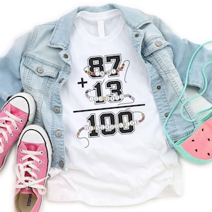 May include: White t-shirt with black text that reads "87 in my 100 days + 13 = 100 of schooler-a". The numbers are surrounded by colorful beads.