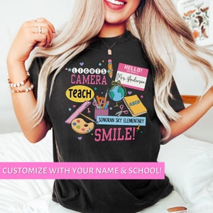 May include: Black t-shirt with a colorful graphic design featuring the text "Lights Camera Teach Smile!" and "Hello! My name is Mrs. Anderson" with a school name and other school-related imagery.