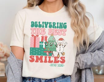 May include: A white t-shirt with a Christmas-themed graphic. The graphic features a smiling Christmas tree and a snowman holding a gift. The text on the shirt reads "Delivering the Best Smiles - Ortho Squad - "