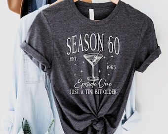 Può includere: Una maglietta grigia scura con testo bianco che recita "SEASON 60 EST. 1965" con una grafica a forma di bicchiere da martini e il testo "Episode One JUST A TINI BIT OLDER".