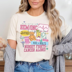 May include: A white t-shirt featuring a pink and blue graphic design that reads "Pediatric Hem/Onc" and "Nobody Fights Cancer Alone!" The design also includes a teddy bear, a stethoscope, a heart rate monitor, and other medical symbols.