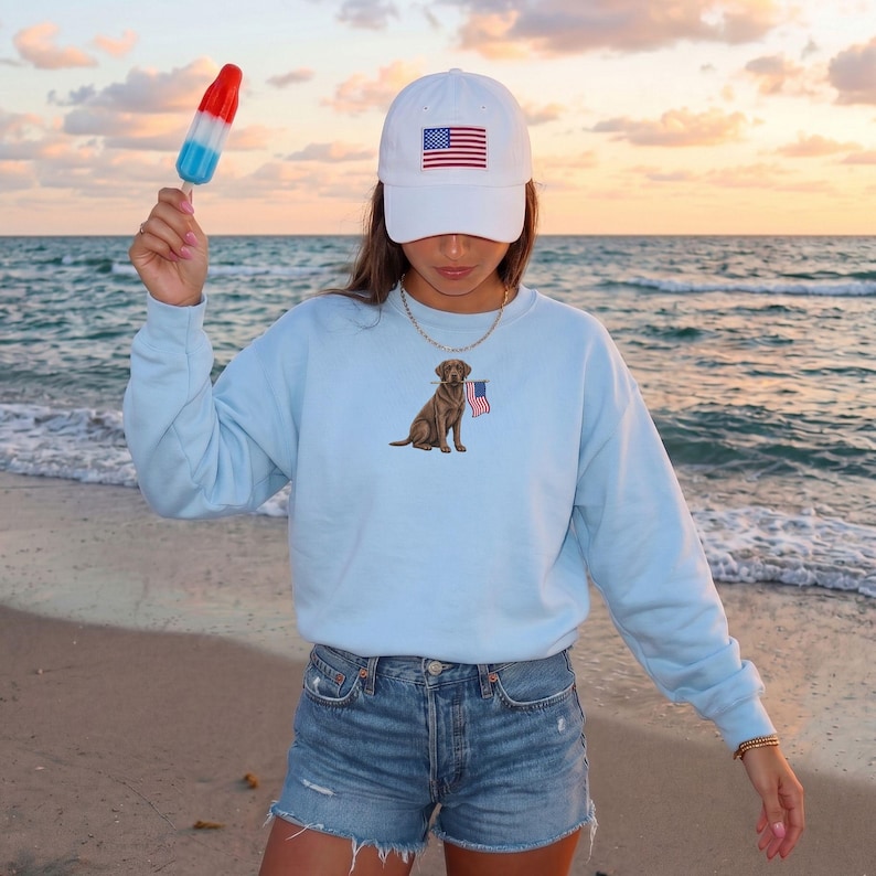May include: A light blue sweatshirt featuring an embroidered brown Labrador Retriever holding a small American flag. The person is wearing a white baseball cap with an American flag patch and denim shorts. A red, white, and blue popsicle is held up.
