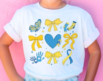 May include: White t-shirt featuring a blue heart, yellow bows, blue socks, a butterfly, and a handprint. The heart is broken and has a blue ribbon on it. The handprint is blue and yellow.