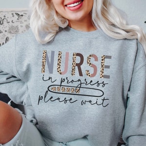 May include: A gray sweatshirt with the text "NURSE in progress please wait" in a leopard print design. The letters are in different colors, including pink, gray, and brown.