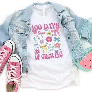 May include: White t-shirt with a pink graphic that says "100 Days of Growing" with colorful flowers and bows.