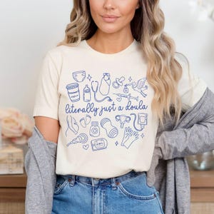 May include: Cream-coloured t-shirt featuring a blue line drawing of baby-related items and the text "literally just a doula". The design includes a baby bottle, stethoscope, and stork.