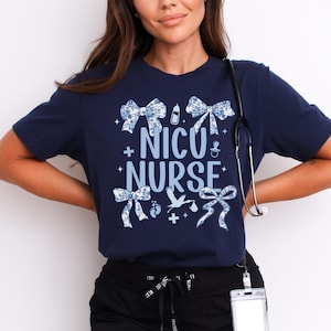 May include: A navy blue t-shirt with the text "NICU NURSE" in white with blue accents. The shirt is decorated with blue bows, a baby bottle, a pacifier, a stork, and footprints.