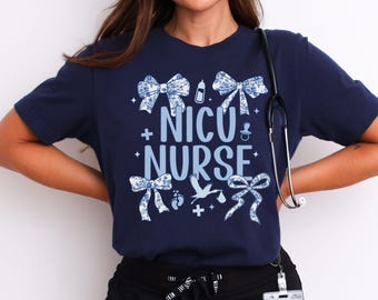 May include: A navy blue t-shirt with the text "NICU NURSE" in white with blue accents. The shirt is decorated with blue bows, a baby bottle, a pacifier, a stork, and footprints.