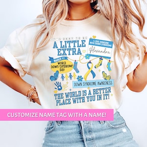 May include: A white t-shirt with a blue and yellow design that says "It's okay to be a little extra" and "Down Syndrome Awareness". The shirt also has a name tag that says "I love someone with Down Syndrome Alexandra".
