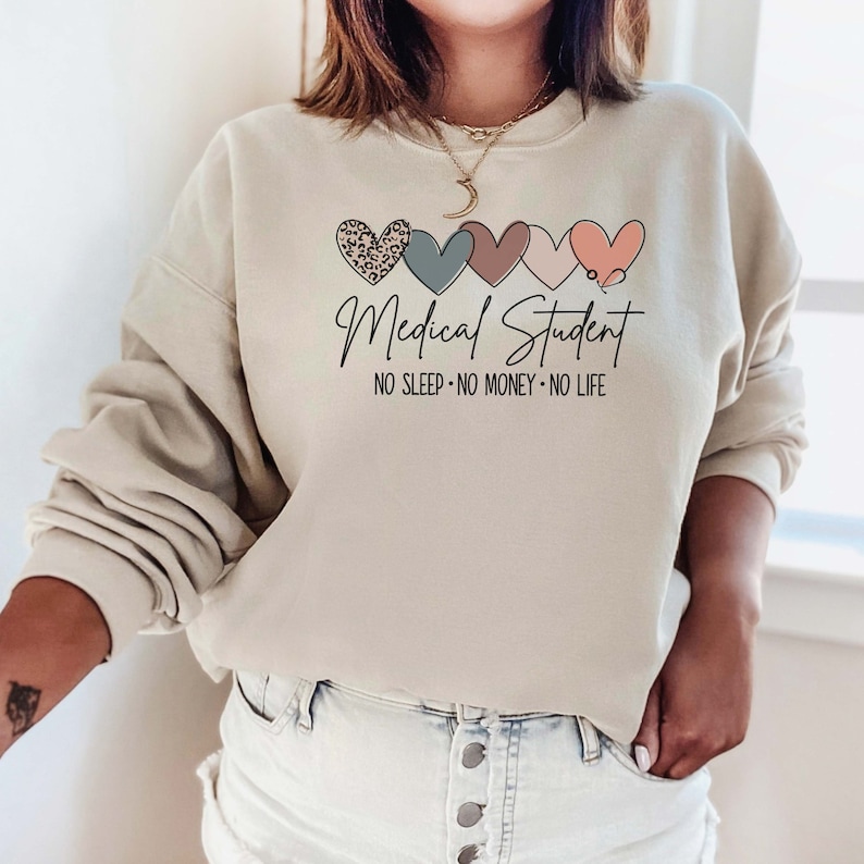 May include: Beige sweatshirt with the text "Medical Student" and the phrase "NO SLEEP - NO MONEY - NO LIFE" in black script. Above the text are five hearts in various colors and patterns, including leopard print and a stethoscope.