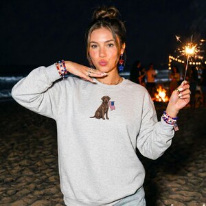 May include: A light gray sweatshirt featuring an embroidered brown dog holding a small American flag. The sweatshirt has long sleeves and a crew neck. The image is taken at night with a beach and bonfire in the background.