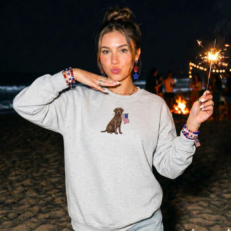 May include: A light gray sweatshirt featuring an embroidered brown dog holding a small American flag. The sweatshirt has long sleeves and a crew neck. The image is taken at night with a beach and bonfire in the background.