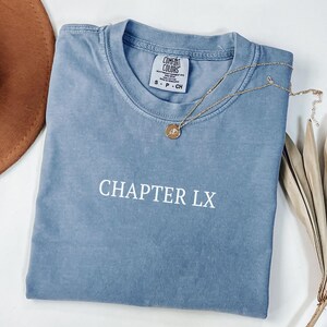 May include: A blue Comfort Colors t-shirt with the text "CHAPTER LX" in white. A gold necklace with a pendant rests on the shirt. The shirt is folded on a white surface.