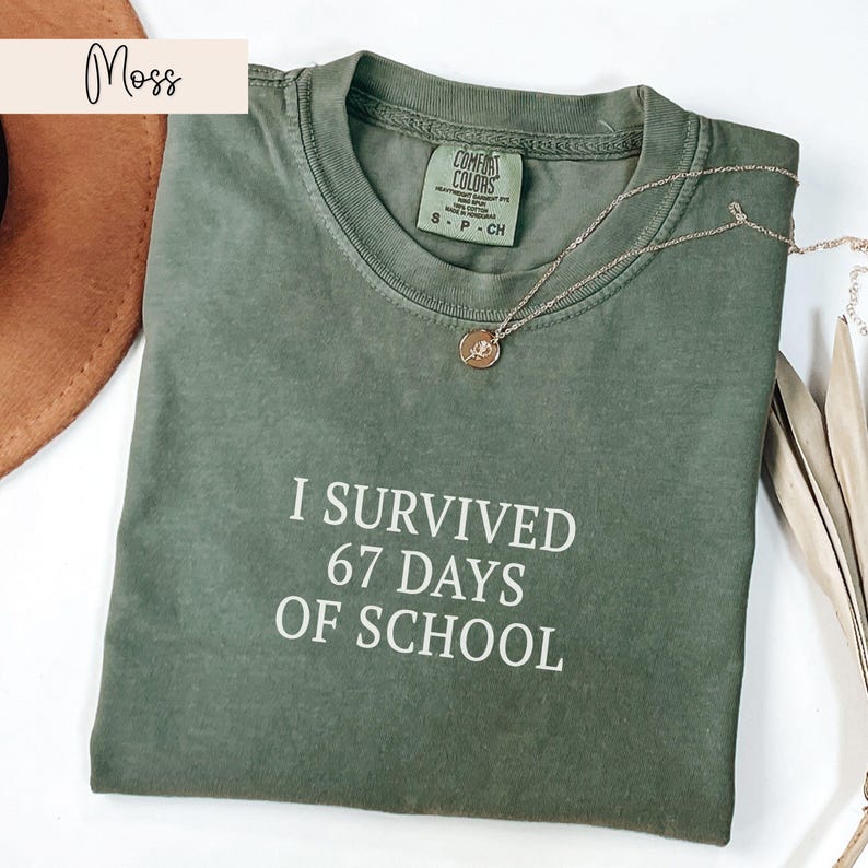 May include: A moss green Comfort Colors t-shirt with the text "I SURVIVED 67 DAYS OF SCHOOL" in white. A gold necklace with a pendant is draped across the shirt. The shirt is a size small.