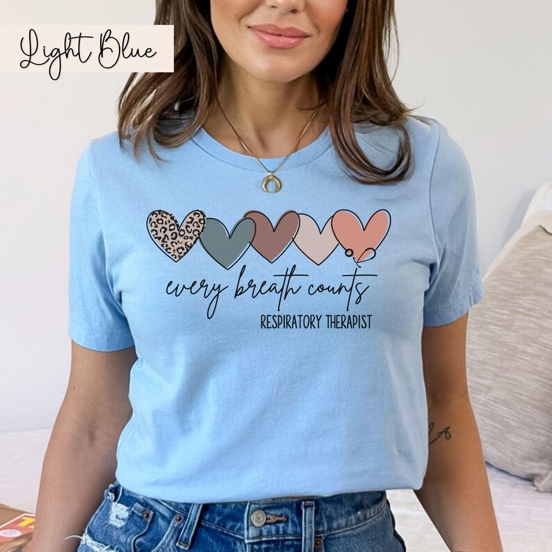 以下が含まれることがあります： 薄い青色のTシャツに、異なる色と模様の5つのハートが並んでいます。ハートは聴診器の線でつながっています。ハートの下には「every breath counts」というテキストがあり、その下には「RESPIRATORY THERAPIST」というテキストがあります。