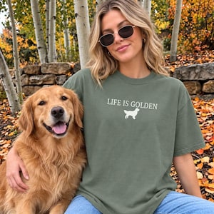 May include: An olive green short-sleeved t-shirt featuring the white text "LIFE IS GOLDEN" centered above a white silhouette graphic of a Golden Retriever dog. The shirt has a crew neckline and a relaxed fit, made of a solid-colored cotton fabric.