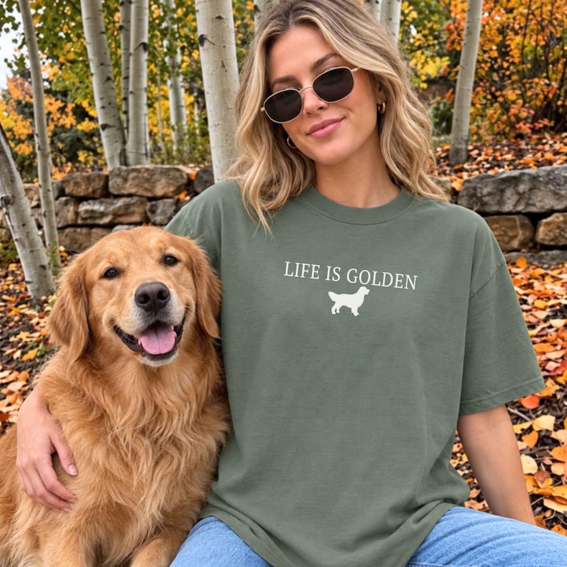 May include: An olive green short-sleeved t-shirt featuring the white text "LIFE IS GOLDEN" centered above a white silhouette graphic of a Golden Retriever dog. The shirt has a crew neckline and a relaxed fit, made of a solid-colored cotton fabric.