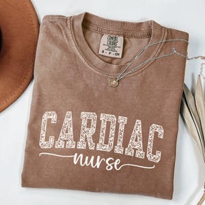 Op de afbeelding: Een bruin t-shirt met de woorden "CARDIAC nurse" in witte letters. Het woord "CARDIAC" is in een decoratief bloemenlettertype. Een zilveren ketting met een kleine hanger is over het shirt gedrapeerd. Het shirt is van het merk Comfort Colors.