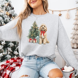 May include: A light gray sweatshirt featuring a watercolor illustration of a Cockapoo dog wearing a red bow, a Christmas tree, and wrapped gifts. The sweatshirt is a comfortable, casual style.