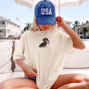Op de afbeelding: Cr&egrave;mekleurig T-shirt met een geborduurde teckel met een Amerikaanse vlag. De hond is zwart en bruin. Een blauwe baseballcap met "USA" in witte letters wordt gedragen. De foto is buiten op een zonnige dag genomen.
