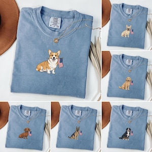 May include: A blue crew neck t-shirt featuring a small embroidered graphic of a dog holding an American flag in its mouth. The shirt has a Comfort Colors label at the inner neckline. Several variations are shown, each displaying a different dog breed including a Corgi, French Bulldog, Golden Retriever, Dachshund, German Shepherd, and Bernese Mountain Dog.