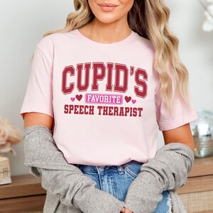 May include: A light pink t-shirt with the text "Cupid's Favorite Speech Therapist" in maroon lettering. The text is arranged in a collegiate style with hearts. The shirt is being worn by a person.