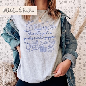 以下が含まれることがあります： グレーヘザーのTシャツに、青いグラフィックデザインが施されています。デザインには「文字通りプロのしゃべり屋」という文字と、マイク、ラップトップ、コーヒーカップなどのアイコンが描かれています。