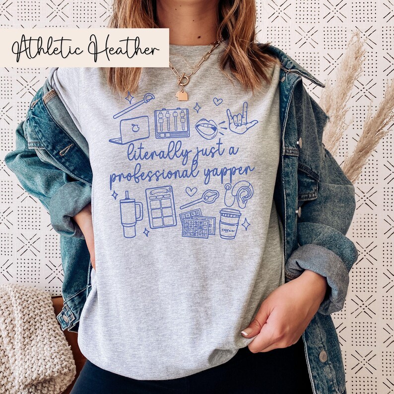以下が含まれることがあります： グレーヘザーのTシャツに、青いグラフィックデザインが施されています。デザインには「文字通りプロのしゃべり屋」という文字と、マイク、ラップトップ、コーヒーカップなどのアイコンが描かれています。