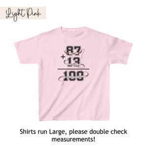 May include: Light pink t-shirt with a black graphic of a math equation: 87 + 13 = 100. The numbers are made of colorful beads.