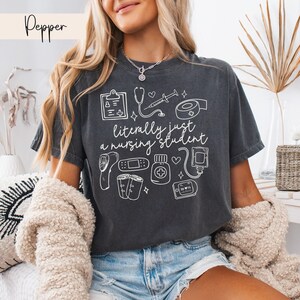 K&ouml;nnte beinhalten: Ein dunkelgraues T-Shirt mit einem wei&szlig;en Grafikdesign von medizinischen Instrumenten und dem Text "literally just a nursing student".