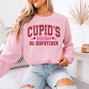May include: A light pink sweatshirt with the text "CUPID'S FAVORITE 911 DISPATCHER" in maroon lettering. The design includes hearts. The sweatshirt is a crew neck style.