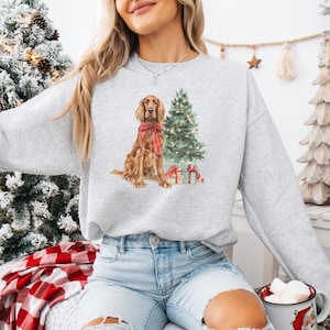 May include: A light gray sweatshirt featuring a watercolor illustration of a brown dog wearing a red bow, a decorated Christmas tree, and wrapped gifts. The sweatshirt is a relaxed fit.
