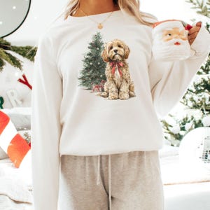 May include: A white sweatshirt featuring a watercolor illustration of a dog with a red bow, a Christmas tree, and wrapped gifts. The sweatshirt is part of a holiday-themed collection.