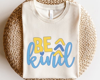 May include: A beige t-shirt with the words "BE kind" in blue and yellow lettering. The letters are stylized with a heart and wavy lines.