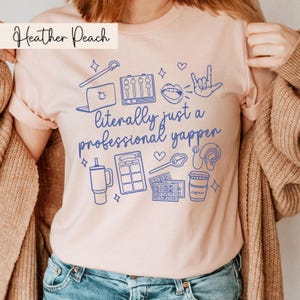以下が含まれることがあります： ピンクのTシャツに、青いグラフィックデザインが施されています。デザインには、「ただのプロのしゃべり屋」という文字が書かれています。グラフィックには、マイク、ラップトップ、コーヒーカップ、ミキシングボード、ハート、愛してるのサインをする手、ヘッドホン、サイコロが描かれています。