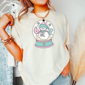 May include: Cream-colored t-shirt featuring a snow globe design with a snowman wearing a pink hat and scarf. The snow globe has the numbers "6" and "7" inside. The base of the snow globe is light blue with a star.