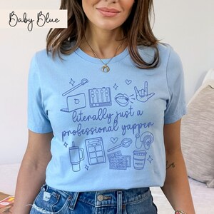 以下が含まれることがあります： ポッドキャスティングに関連するさまざまなアイコン（マイク、ヘッドホン、ラップトップ、コーヒーカップなど）が描かれたライトブルーのTシャツ。Tシャツには「literally just a professional yapper」というテキストがプリントされています。