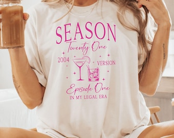 Può includere: Maglietta bianca con testo rosa che recita "SEASON Twenty One 2004 VERSION Episode One IN MY LEGAL ERA". Il testo è circondato da piccole stelle rosa. Ci sono due illustrazioni rosa di cocktail.