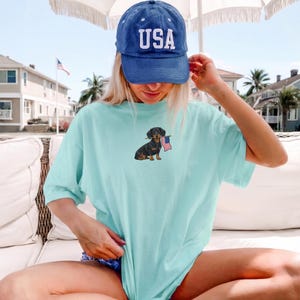 Op de afbeelding: Lichtblauw T-shirt met een teckel die een Amerikaanse vlag vasthoudt. De persoon draagt een blauwe baseballcap met "USA" erop. De afbeelding heeft een casual, patriottisch thema, geschikt voor dagelijks gebruik.