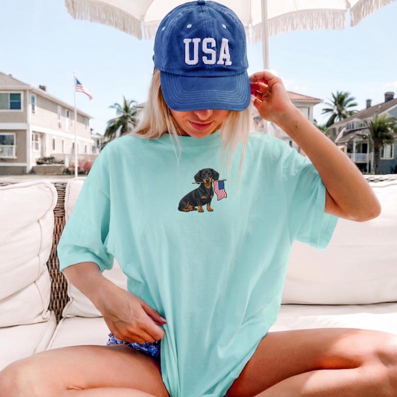Op de afbeelding: Lichtblauw T-shirt met een teckel die een Amerikaanse vlag vasthoudt. De persoon draagt een blauwe baseballcap met "USA" erop. De afbeelding heeft een casual, patriottisch thema, geschikt voor dagelijks gebruik.