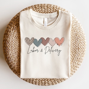 Op de afbeelding: Een beige T-shirt met een rij van vijf kleurrijke harten, elk met een ander patroon. De harten zijn zwart omrand en het laatste hart heeft een stethoscoop. De tekst "Labor & Delivery" staat onder de harten.